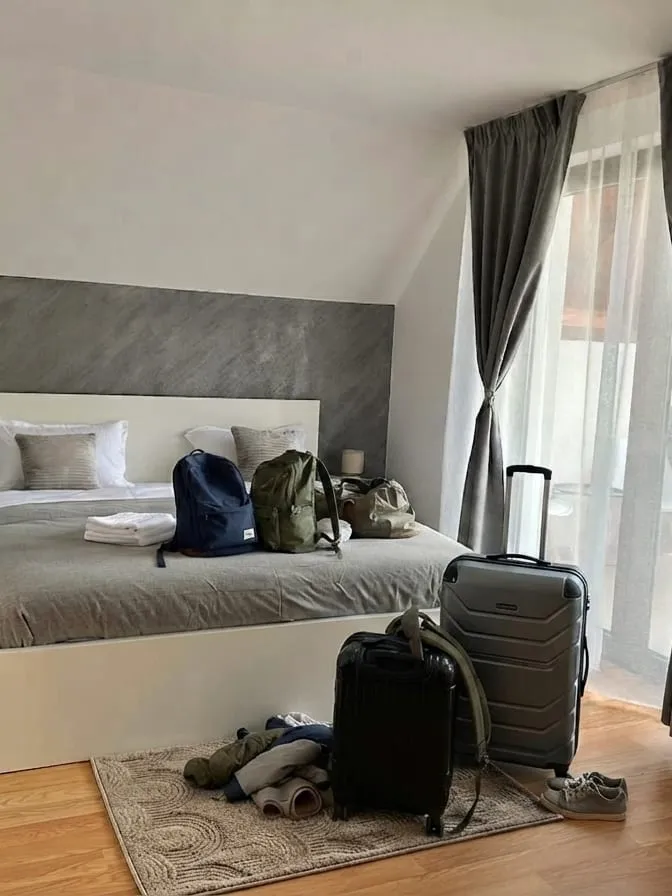 Același dormitor la sosire: rucsacuri și o geantă de voiaj pe pat, două valize și o pereche de pantofi pe covorul de la picioarele patului, cu draperiile trase spre balcon
